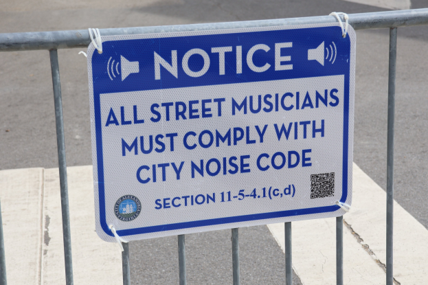 Anschlagtafel an einem Zaun mit einem Schild, das Straßenmusikanten zur Einhaltung städtischer Lärmvorschriften auffordert, mit einer Straße im Hintergrund.