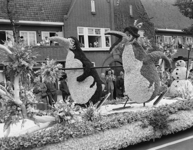 Schwarz-weiß-Foto eines Paradewagens mit Blumen und Pinguinfiguren, umgeben von Zuschauern, Bäumen und Gebäuden.