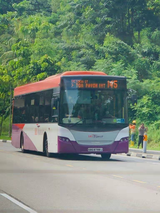 Ein lila und weißer SBS Transit Bus fährt eine von Bäumen gesäumte Straße entlang mit ein paar Passagieren drinnen und zwei Menschen, die auf dem Bürgersteig stehen.