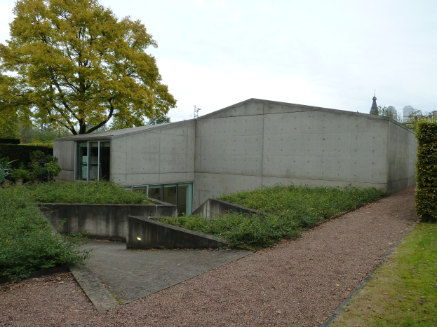 Concrete building with a walkway, surrounded by green grass and plants, trees in the background, and a clear blue sky.