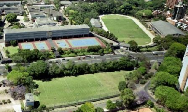 Luftaufnahme eines Fußballfeldes auf dem Campus der University of Sydney, umgeben von Bäumen, Gebäuden und Fahrzeugen, mit einem neuen Sportkomplex im Bau.