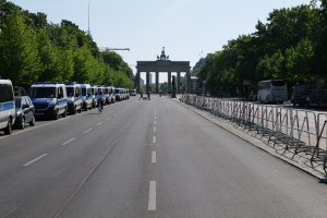 Eine Reihe von Polizeiwagen, die auf der Seite einer Straße vor dem Brandenburger Tor in Berlin, Deutschland, geparkt sind, mit Menschen, die Fahrräder fahren und auf der Straße stehen, Absperrungen, Bäumen und einem Bogen mit Statuen im Hintergrund.