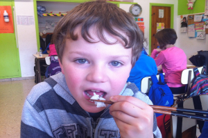 Ein Junge in einer grauen und schwarzen Jacke isst mit Stäbchen in einem Klassenzimmer, mit anderen Kindern hinter ihm, einer grünen und weißen Wand, Lehrplakaten und Diagrammen an einer weißen Wand, einer braunen Tür und einer Wanduhr.