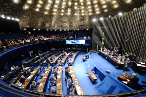 Eine Konferenzb├╝hne mit sitzenden Personen an einem Tisch mit Papieren und Gegenst├Ąnden, flankiert von Flaggen und einer Skulptur, mit Zuschauern an Tischen und hinter einer Absperrung, beleuchtet von Deckenleuchten.