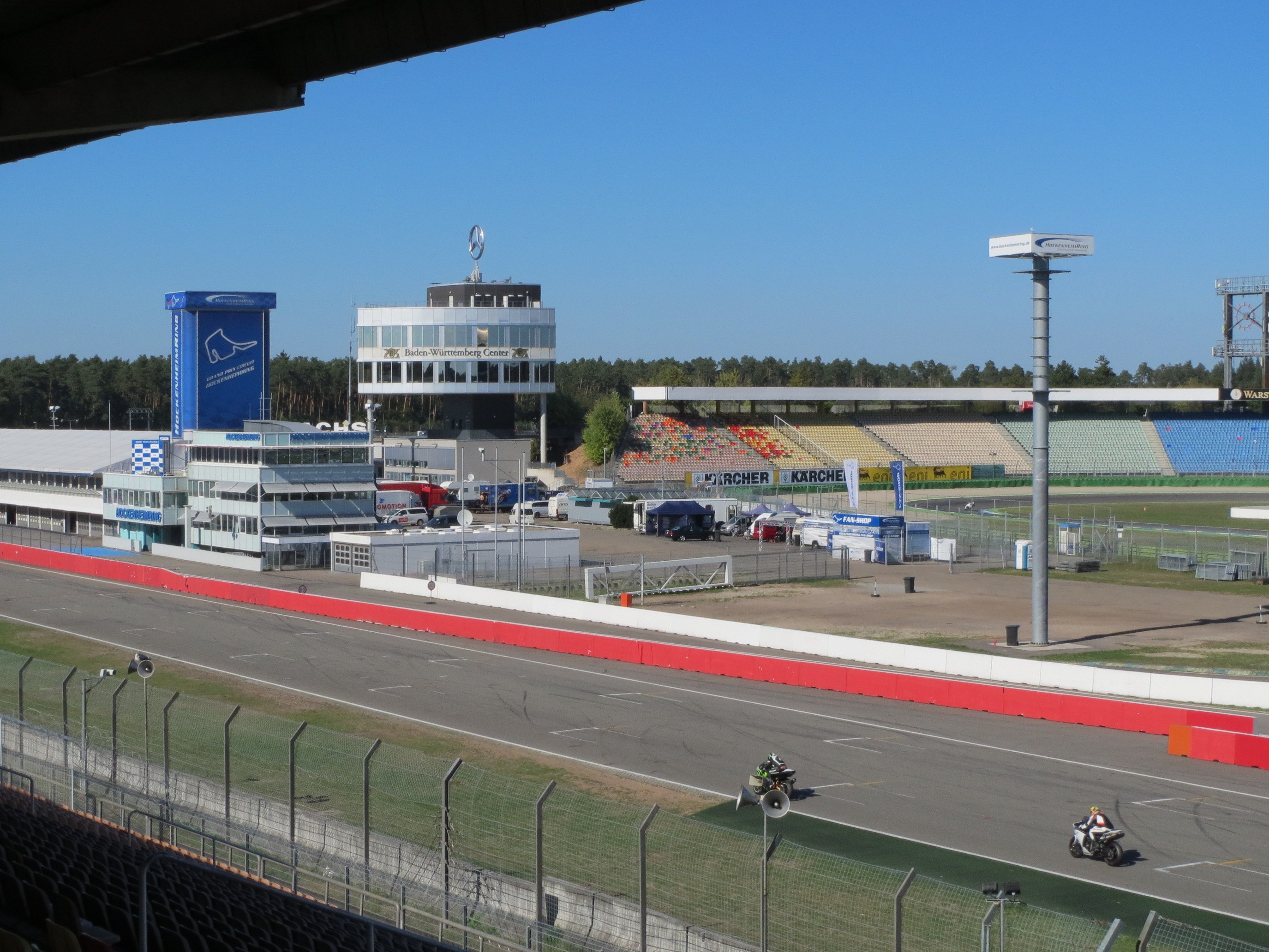 Gruppe von Motorradfahrern auf einer Rennstrecke, umgeben von einem Metallzaun, Gras und einem bewölkten Himmel.