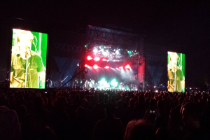 Eine Konzertszene mit einer Menschenmenge unten, einem Bildschirm links und einem weiteren Bildschirm rechts.