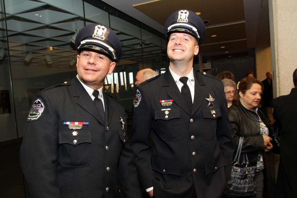Zwei grinsende Beamte im Vordergrund, mit Menschen in der Mitte, Glasfenster links, Lampen und Decke oben und Säulen und Menschen rechts.