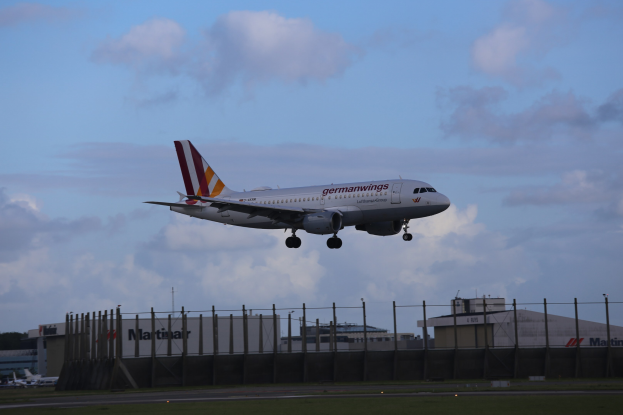 Germanwings Airbus A320-200 beim Starten am Frankfurter Flughafen, mit Gras darunter, einem Zaun im Vordergrund, Gebäuden und Bäumen im Hintergrund und einer wolkigen Wolkendecke.