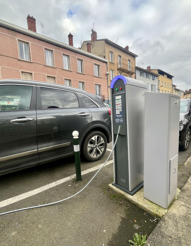 Elektroauto-Laden an einer Straßenstation mit Gebäuden, Laternenmasten und bewölktem Himmel im Hintergrund.