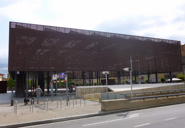 Großes modernes Gebäude mit zahlreichen Fenstern, das als neuer städtischer Eingang dient, flankiert von Straßeninfrastruktur, Fahrzeugen, Fußgängern und Bäumen unter einem bewölkten Himmel.
