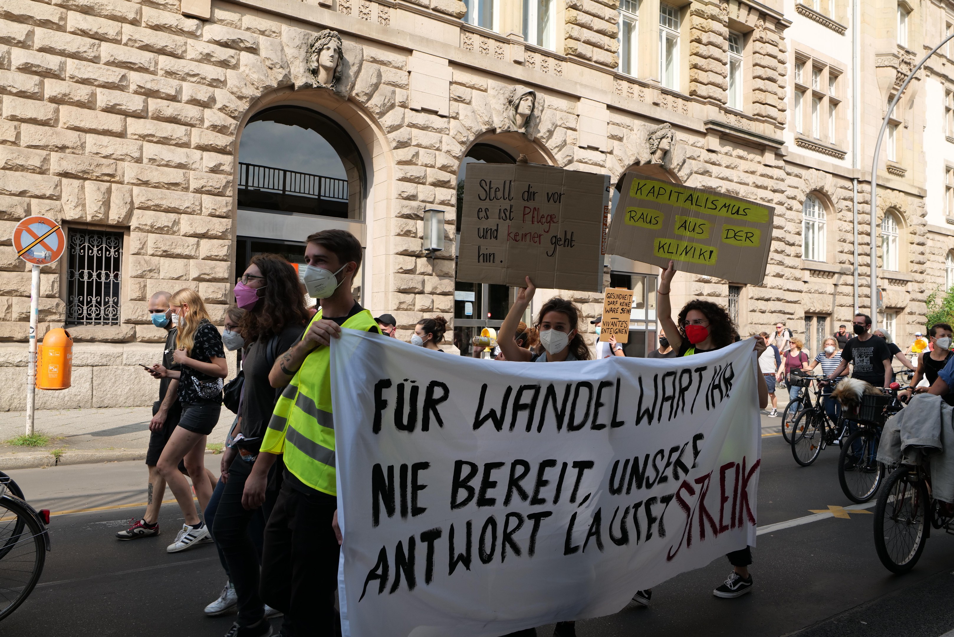 Eine Gruppe von Menschen marschiert auf einer Straße bei einer Demonstration, einige halten Schilder und andere fahren Fahrräder, im Hintergrund ein Gebäude mit Fenstern, Bögen, Säulen und Skulpturen.
