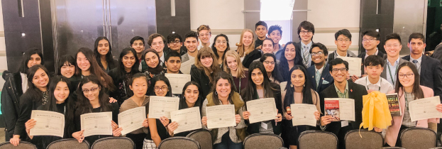 Eine Gruppe von Menschen posiert mit Urkunden vor einem Hintergrund aus Stühlen und einer Wand, wahrscheinlich bei einer Abschlussfeier 2019-2020.