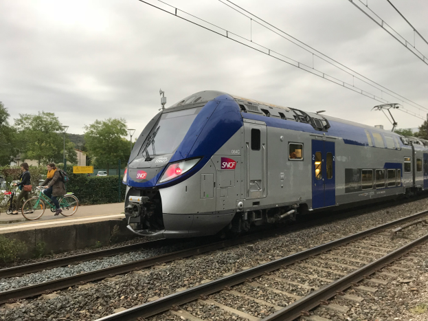 Ein blauer und silberner Zug fährt auf Schienen neben einem Bahnsteig mit Fahrradfahrern, Bäumen, Pfählen, Drähten und einem klaren blauen Himmel im Hintergrund.