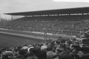 Schwarzes und weißes Foto eines vollen Stadions mit Zuschauern, die ein Fußballspiel beobachten, einige stehen und einige sitzen, mit Bannern, Pfosten, einer Hütte, Bäumen, einem Turm und einem bewölkten Himmel im Hintergrund.