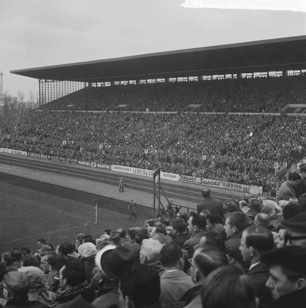 Schwarzes und weißes Foto eines vollen Stadions mit Zuschauern, die ein Fußballspiel beobachten, einige stehen und einige sitzen, mit Bannern, Pfosten, einer Hütte, Bäumen, einem Turm und einem bewölkten Himmel im Hintergrund.