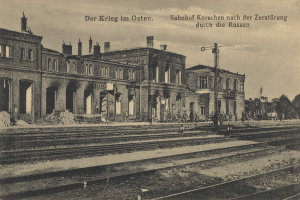 Schwarzes und weißes Foto des Bahnhof Korschen in Ostern, Deutschland, mit Eisenbahnschienen, Gebäuden, Pfählen, Menschen, Bäumen und Himmel mit Text oben und unten.