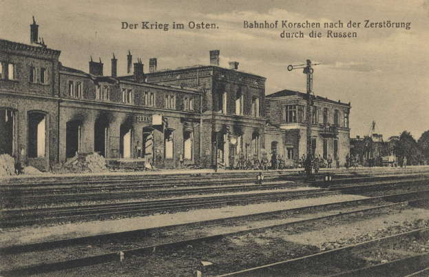 Schwarzes und weißes Foto des Bahnhof Korschen in Ostern, Deutschland, mit Eisenbahnschienen, Gebäuden, Pfählen, Menschen, Bäumen und Himmel mit Text oben und unten.