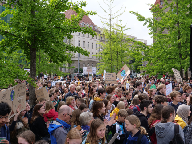Große Menge von Menschen protestiert vor einem Gebäude in Berlin, hält Plakate, mit Bäumen, Fahrzeugen, einem Lautsprecher und Himmel.