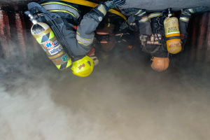 Eine Gruppe von Feuerwehrleuten in Helmen, Handschuhen und Sauerstoffflaschen arbeitet daran, ein Feuer in einem Tunnel zu löschen, wobei Rauch aus der Unterseite aufsteigt.