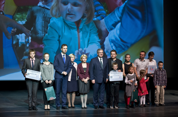 Gruppe von Menschen auf einer Bühne mit Taschen und Urkunden, im Hintergrund ein Bildschirm mit einem Kind und anderen Personen.