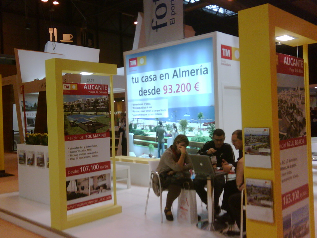 Eine Gruppe von Menschen, die um einen Tisch herum auf Stühlen sitzen und auf einen Laptop schauen und arbeiten, mit mehreren Flyern, auf denen verschiedene Werbeanzeigen zu sehen sind.