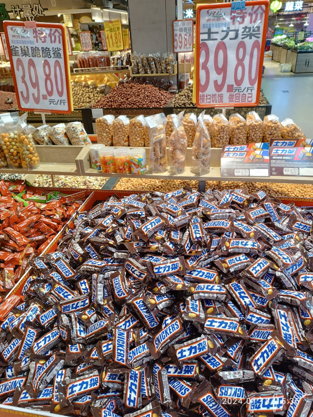 Vitrine mit verschiedenen Süßigkeiten, einschließlich Snickers, in einem Geschäft mit Lebensmitteln, nummerierten Schildern, Deckenleuchten und Hintergrundobjekten.