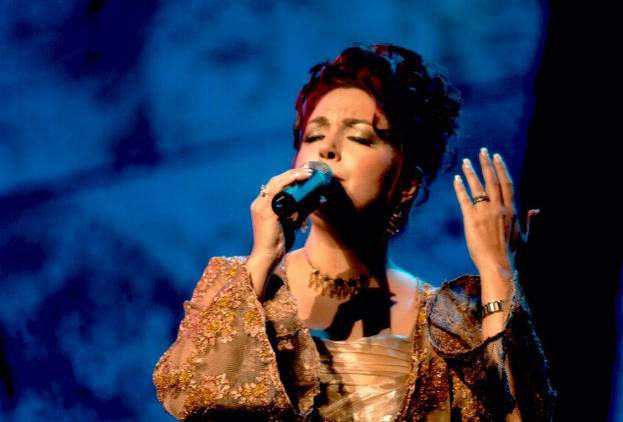 Frau in goldenem Kleid, die auf der Bühne in ein Mikrofon singt, vor blau-schwarzem Hintergrund.