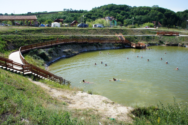 Gruppe von Menschen, die in einem Gewässer mit üppiger grüner Umgebung, einer Brücke mit Stufen zum Wasser, Hütten, Fahrzeugen, Pfählen und einem klaren blauen Himmel im Hintergrund schwimmen.