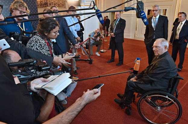 Ein Mann im Rollstuhl umgeben von Reportern mit Kameras, Mikrofonen und Mobiltelefonen sowie einem Banner, Scheinwerfern und einer Wand im Hintergrund.