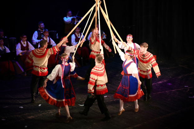 Eine Gruppe von Menschen in bunten Kostümen tanzt um einen Maibaum, einige halten Holzstöcke, während Musiker im Hintergrund Instrumente spielen.