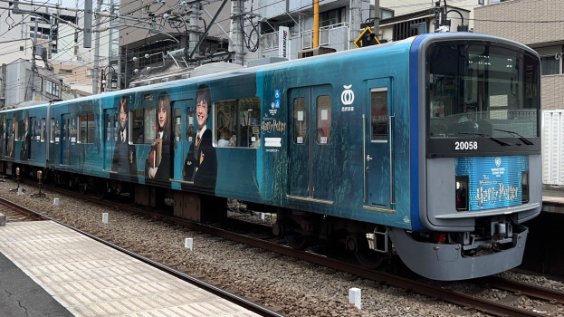 Blauer Zug fährt auf Schienen neben einem Bahnsteig mit Strommasten und Drähten, im Hintergrund Gebäude und einen klaren Himmel.