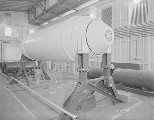 Ein Schwarz-Weiß-Foto einer großen zylindrischen Wasserstoffbombe auf einem Ständer in einer Fabrik, mit Rohren, Lampen und einer Wand im Hintergrund und Text unten.