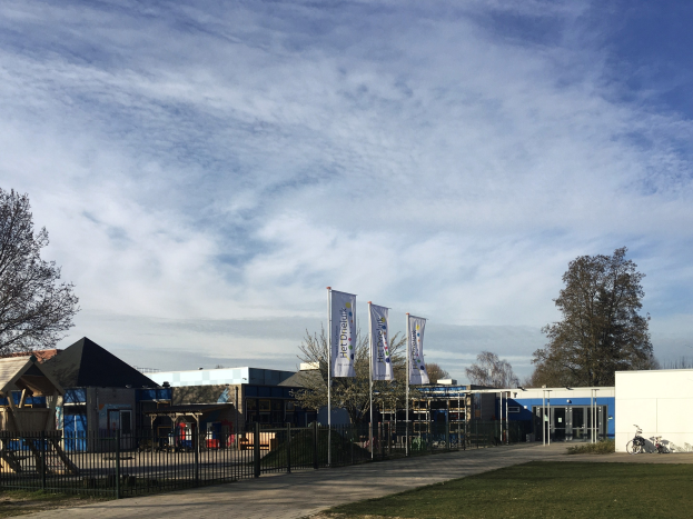 Große Anlage mit einem grünen Bereich vorne, umgeben von einem Metallzaun, mit einem Weg, Bäumen, Masten, Bannern, einem geparkten Fahrrad, näheren Häusern und einem bewölktem Himmel, als der neue Spielplatz der Schule identifiziert.