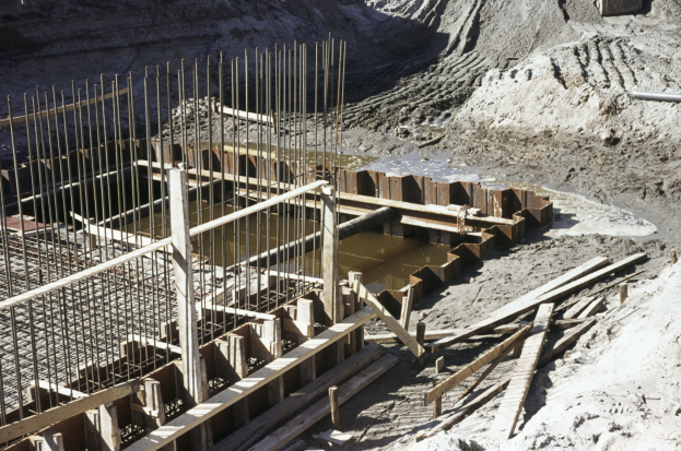 Eine Baustelle mit einem großen Haufen Erde, Holzstöcken, Eisenstangen und Wasser, mit einem Hügel im Hintergrund.