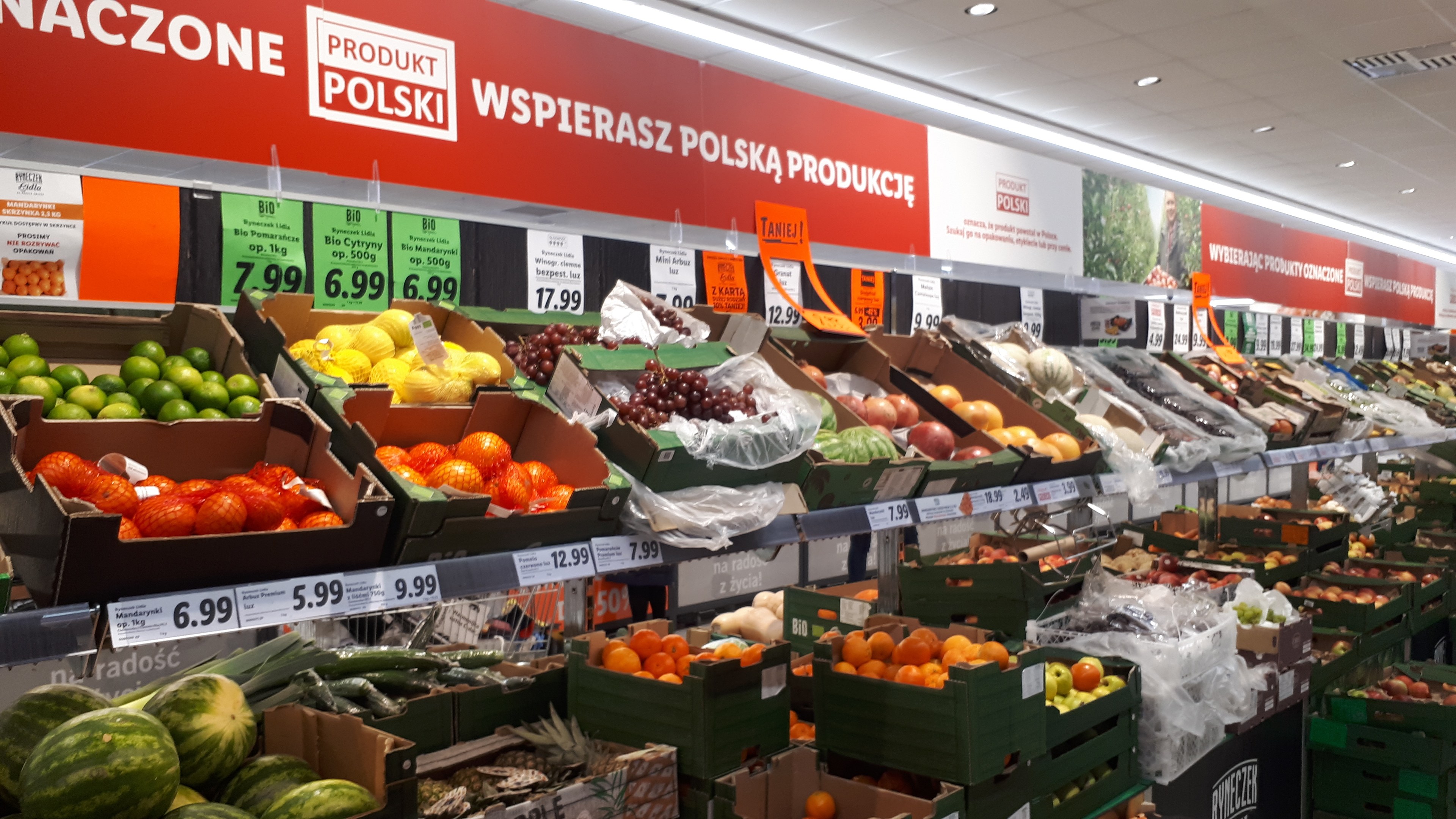 Ein Geschäft mit verschiedenen Obst- und Gemüsesorten in Kartonschachteln, jede mit Preisschildern, beleuchtet von Deckenlampen und mit Informationsschildern an den Wänden.