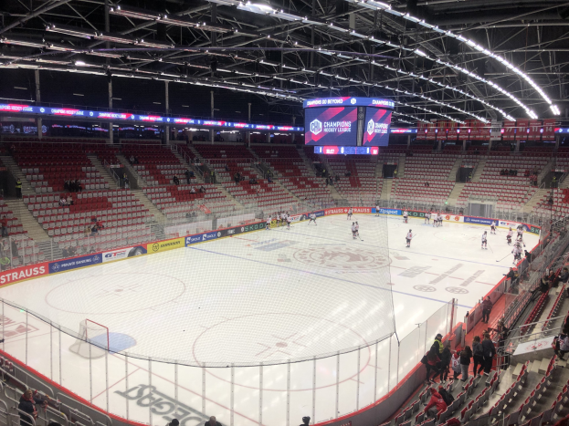 Eishockeyspiel in einer großen Arena mit Zuschauern, eine Eisfläche umgeben von einem Zaun, ein Display mit der Aufschrift "St. Louis Blues vs Detroit Red Wings", Metallrahmen, Deckenleuchten und ein Dach.